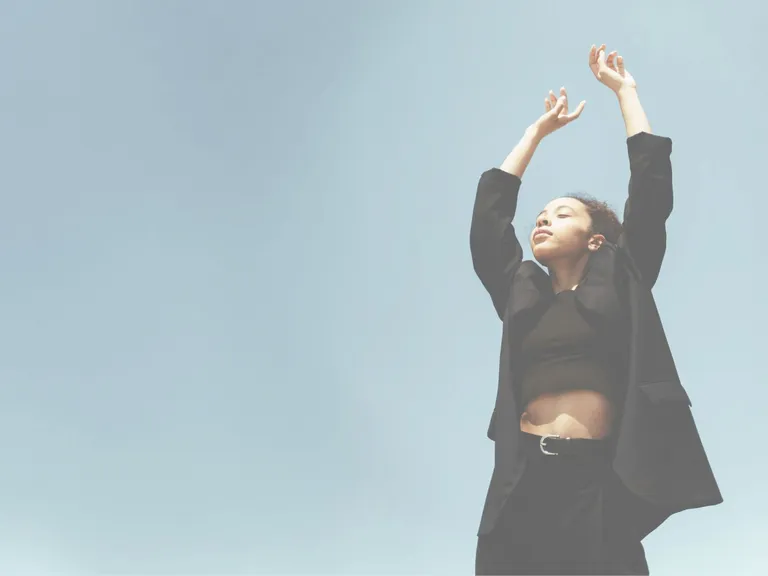 Woman in a black outfit standing with her arms raised against a clear blue sky. Symbolic image representing ellexx invest and long-term ETF investing.